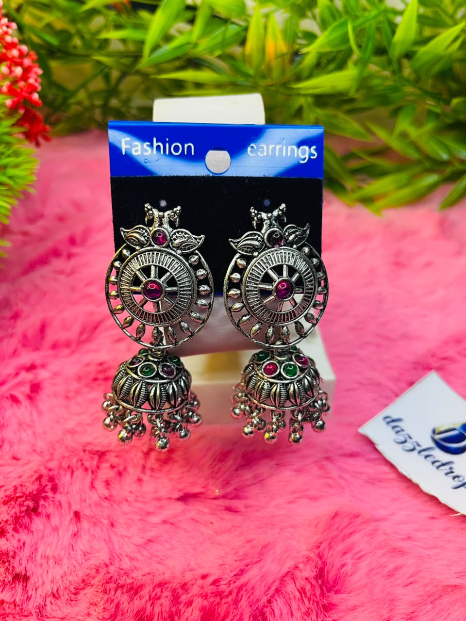 Oxidized silver jhumka earrings featuring ruby red and emerald green stone accents on a pink plush background.
